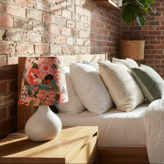 Floral lampshade on a white base next to a bed with pillows against a brick wall.