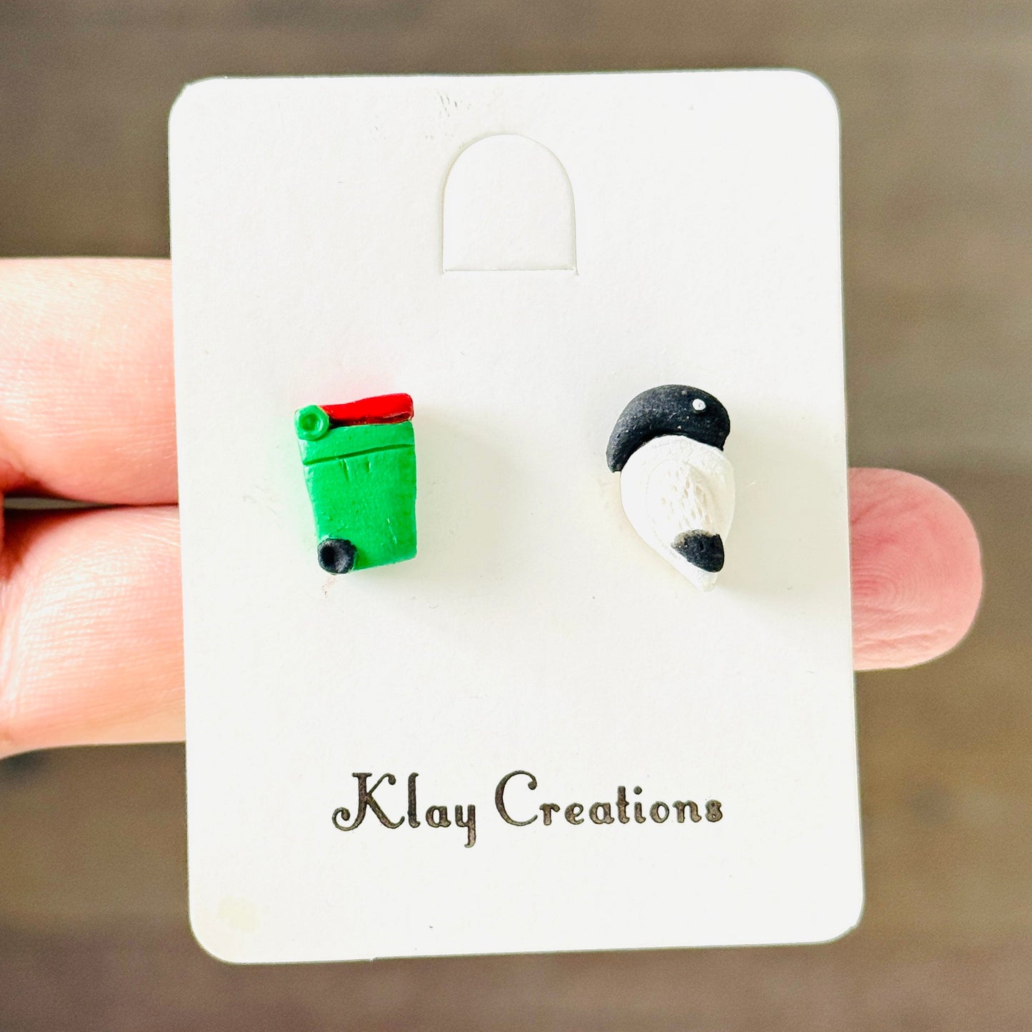 A hand holding a white display card featuring two small clay earrings. One earring is a green trash can with a red lid, and the other is a black-and-white cat head.