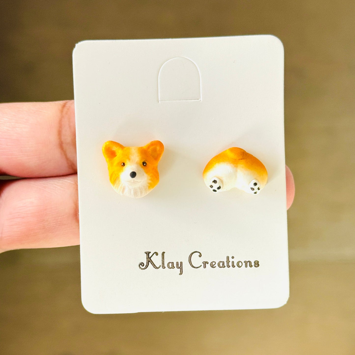 A pair of small corgi-shaped stud earrings on a white display card held by a hand. The card is branded 'Klay Creations' and is photographed against a warm wooden background.