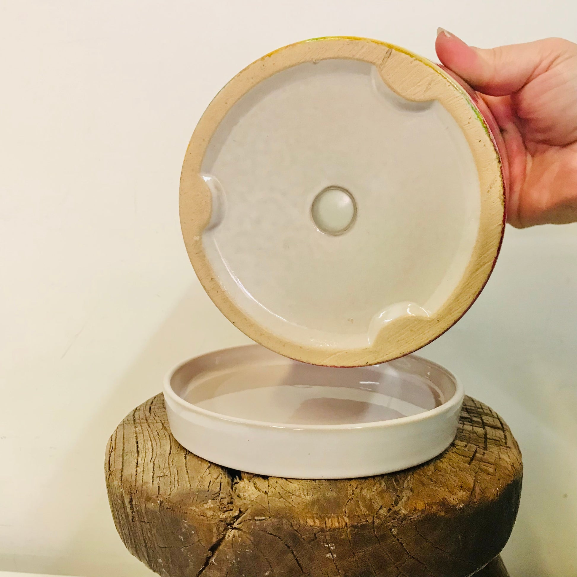 Hand holding a ceramic lid over a container on a wooden block with a white background