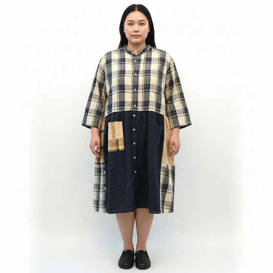 A patchwork button-up shirt dress hanging on a wooden hanger against a plain white wall. The dress features a beige and navy plaid pattern on the upper portion and sleeves, a solid navy quilted lower section, and tan patchwork details on the front pocket and side panels.
