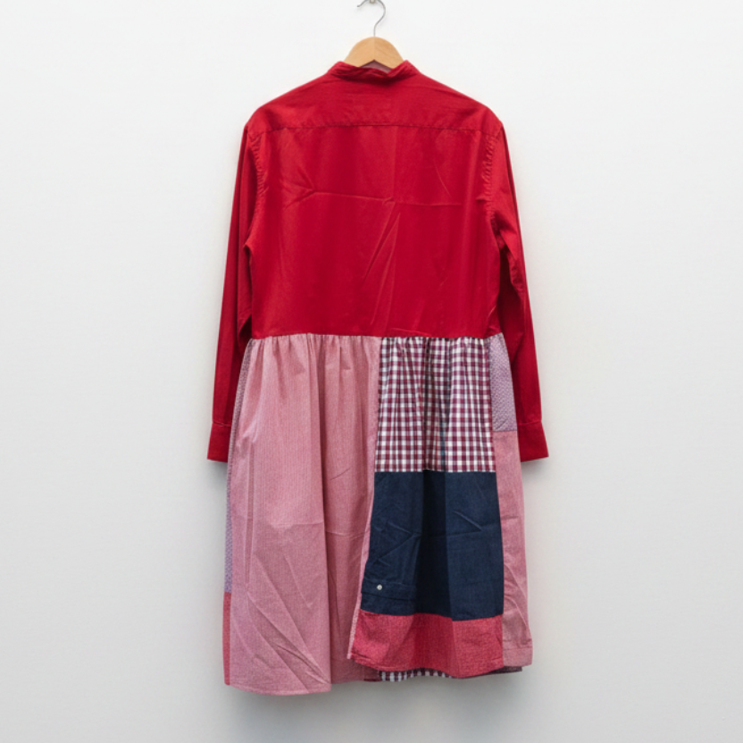 A long-sleeve patchwork dress hanging on a wooden hanger against a white painted brick wall. The dress has a solid red upper bodice and sleeves, a pink gathered waist, and patchwork lower panels in red-and-white gingham, navy blue, and pink.