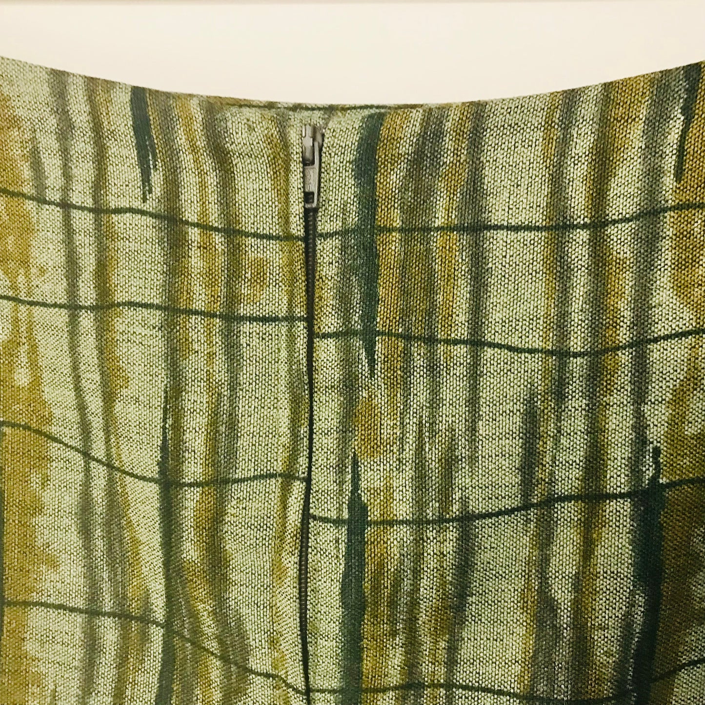 Close-up of a textured woven fabric with a vertical zipper, featuring an abstract pattern of olive green, mustard yellow, and dark charcoal gray vertical streaks over a light beige base.