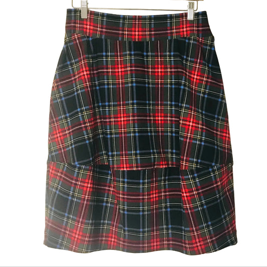 A pleated A-line skirt in a red, black, and blue tartan plaid pattern, hanging from a wooden hanger against a plain off-white wall.