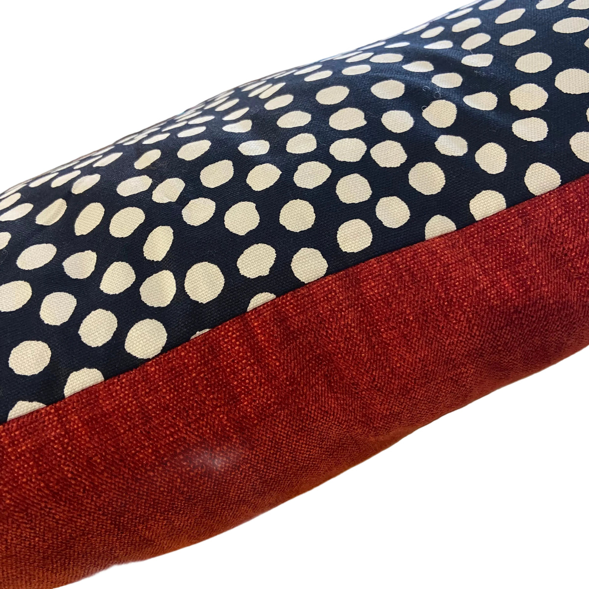 A close-up of a fabric cushion or pillow showing a navy blue top section with cream polka dots and a contrasting rust-red bottom band, photographed in a real indoor setting with shelves blurred in the background.