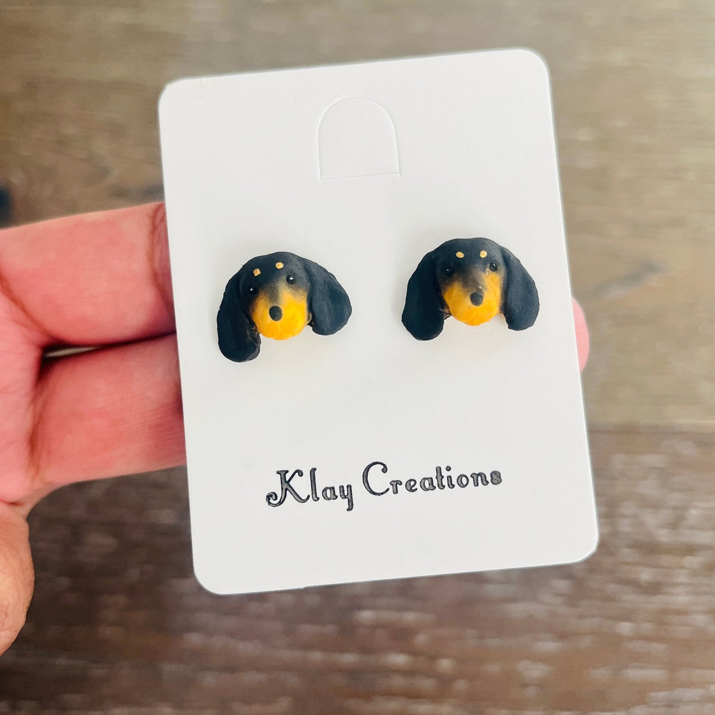 A pair of dachshund-shaped stud earrings on a white display card held by a hand over a wooden surface. The card is branded 'Klay Creations'.