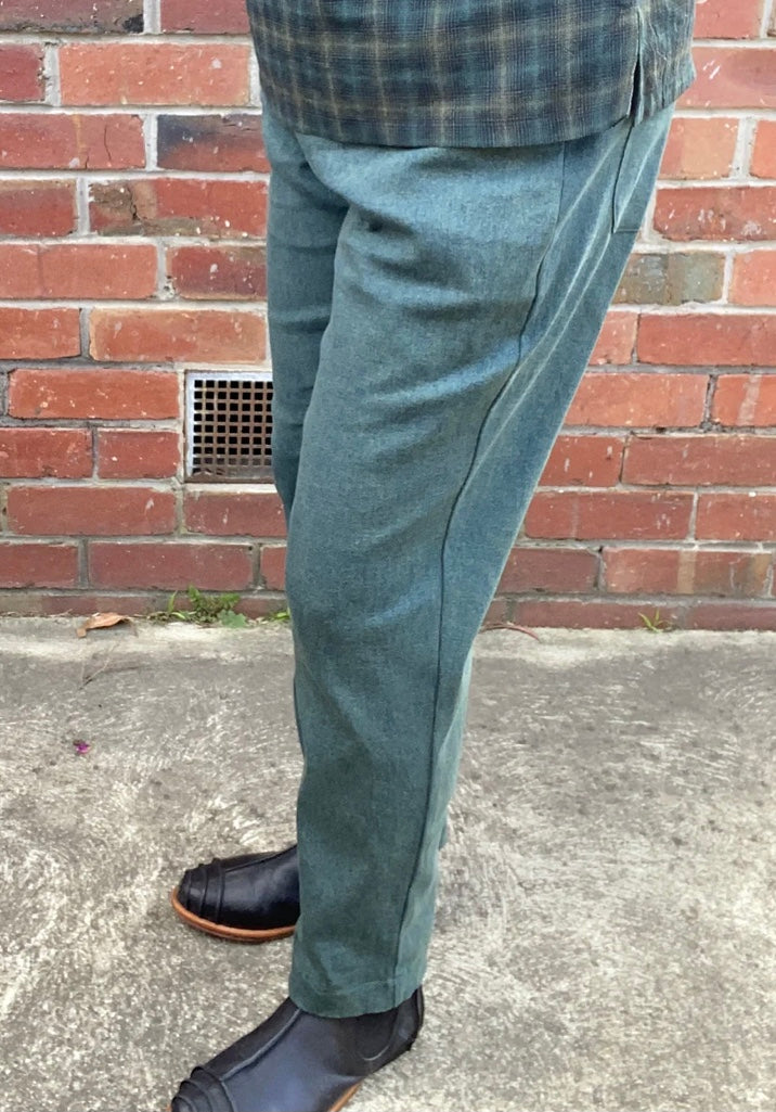 Person wearing green pants and black shoes standing against a brick wall.