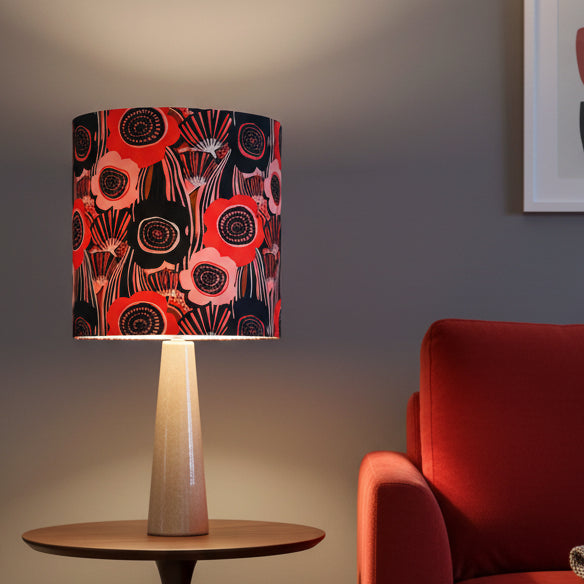 A table lamp with a cylindrical drum shade featuring a bold poppy pattern in red, pink, black, and white, mounted on a tapered pinky bronze base 