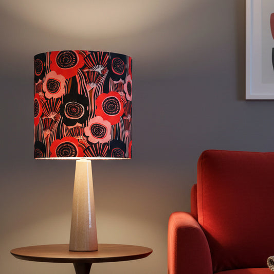 A table lamp with a cylindrical drum shade featuring a bold poppy pattern in red, pink, black, and white, mounted on a tapered pinky bronze base 