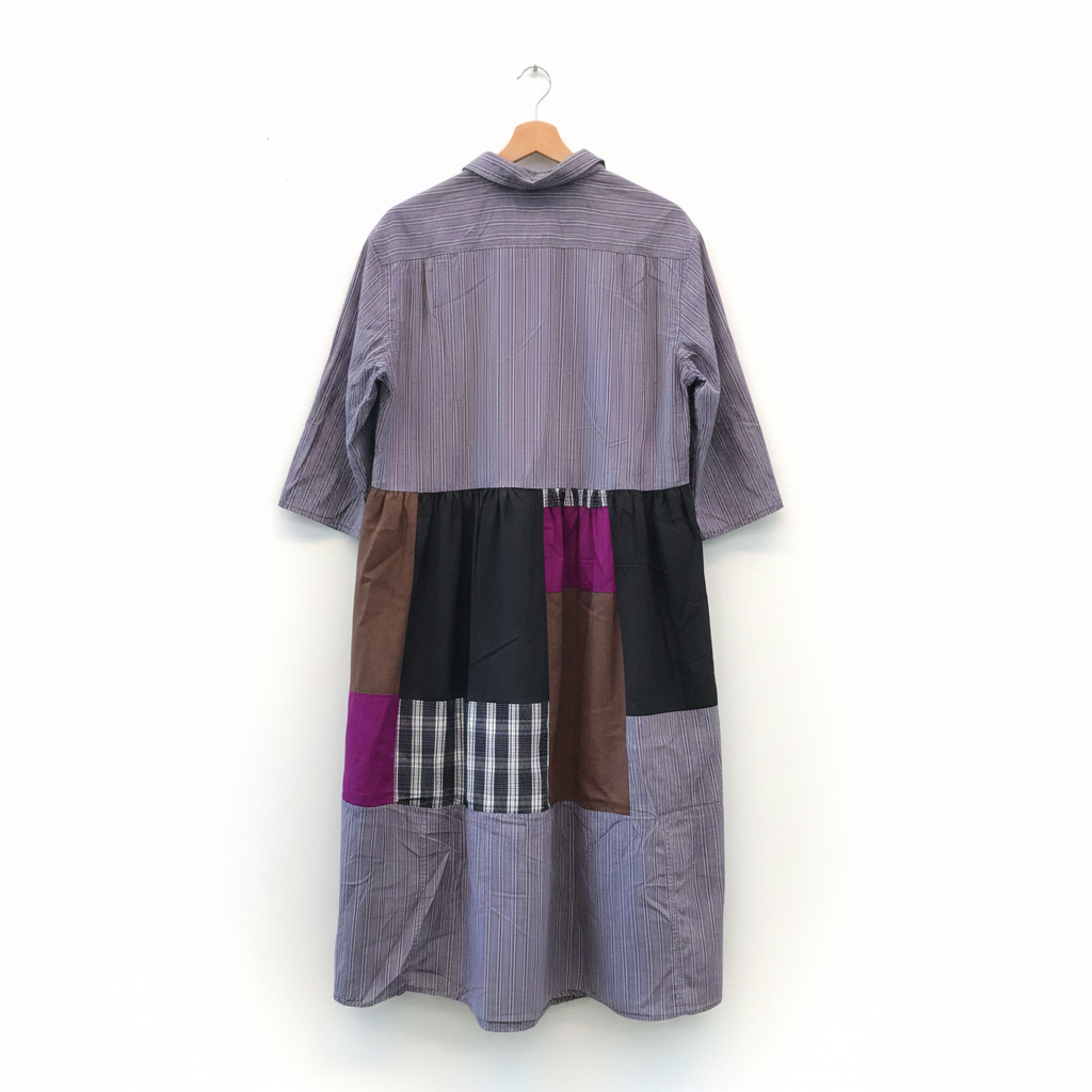 A handmade patchwork shirt dress hanging on a wooden hanger against a white brick wall. The dress features a lavender-gray vertically striped long-sleeve top and a patchwork skirt with sections of black, brown suede-like fabric, purple, plaid, and striped fabrics.