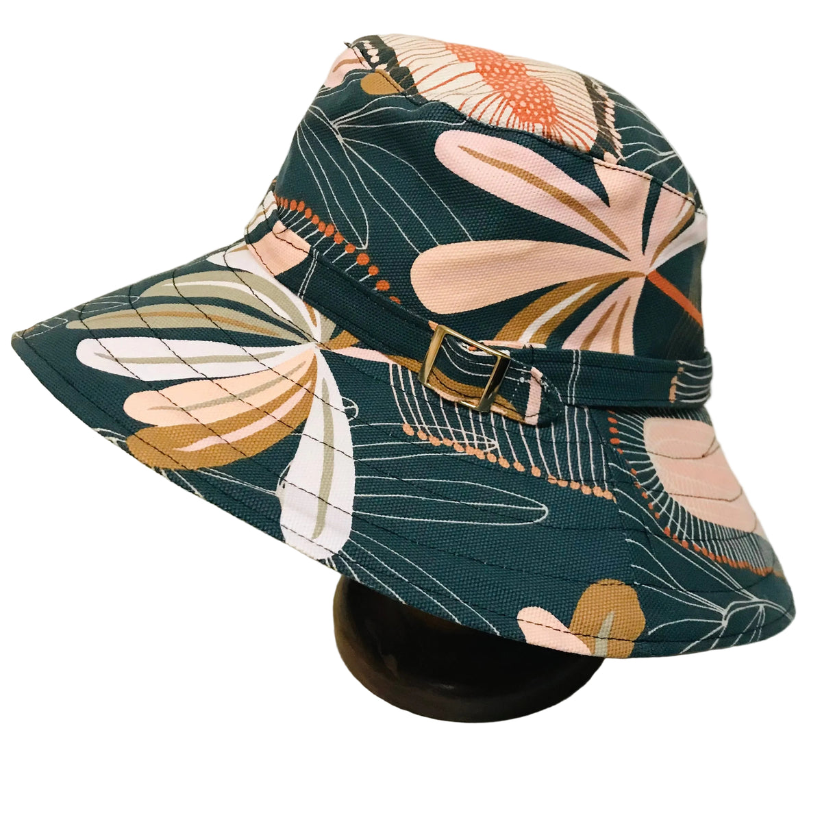 A wide-brimmed bucket hat with a dark teal background and a retro floral pattern in pink, orange, beige, and white. The hat features a matching fabric band with a gold-tone buckle, displayed on a dark stand against a plain off-white background.