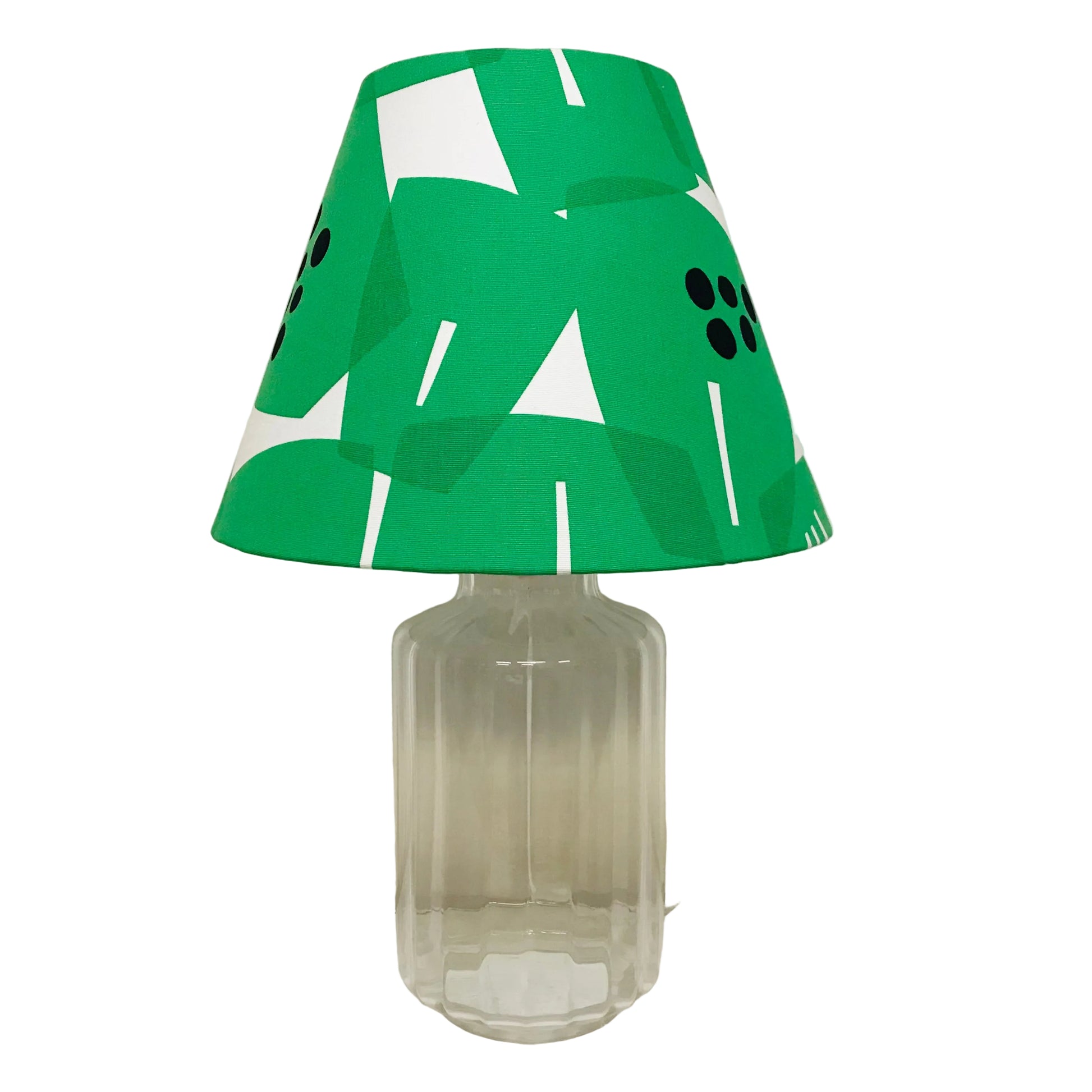 A table lamp with a green, white, and black geometric patterned fabric shade on a clear glass base, set against a plain off-white wall.