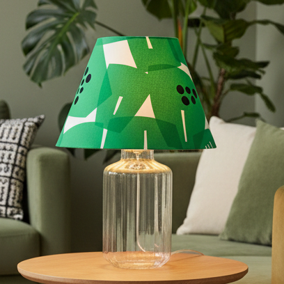 A table lamp with a green, white, and black geometric patterned fabric shade on a clear glass base, set against a plain off-white wall.