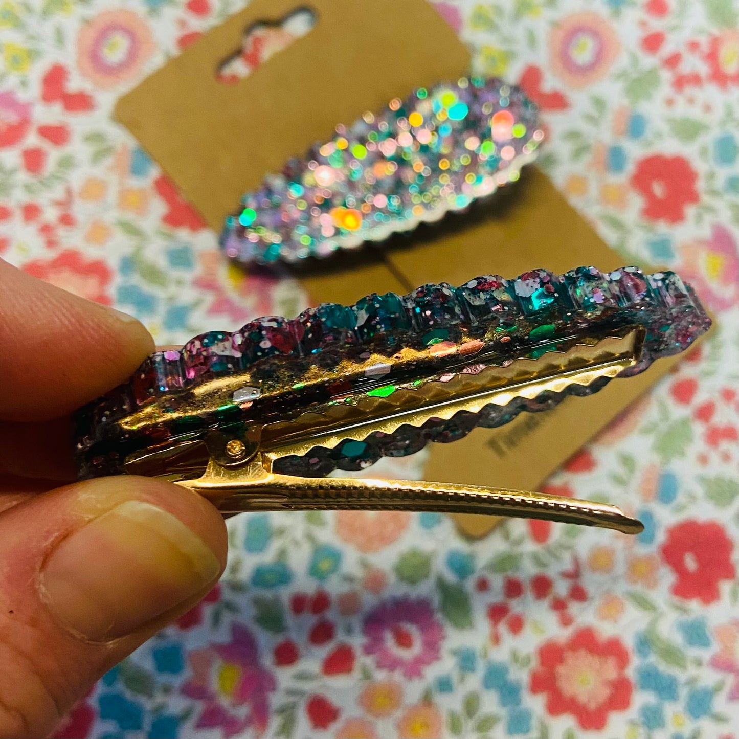 Multicolored glittery hair clip held by a hand with a floral-patterned background