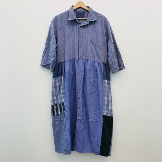 A patchwork button-up shirt dress in shades of purple, blue, and plaid, hanging on a wooden hanger against a white painted brick wall.