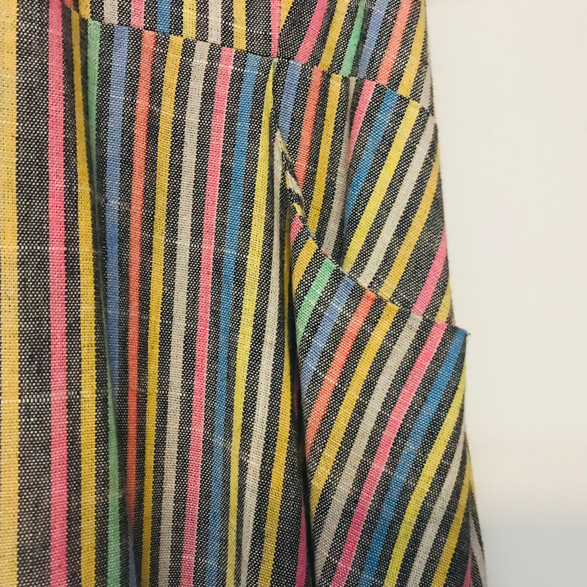 Close-up of a striped woven textile with vertical multicolor lines including yellow, pink, blue, green, black, and white, shown against a plain light background.
