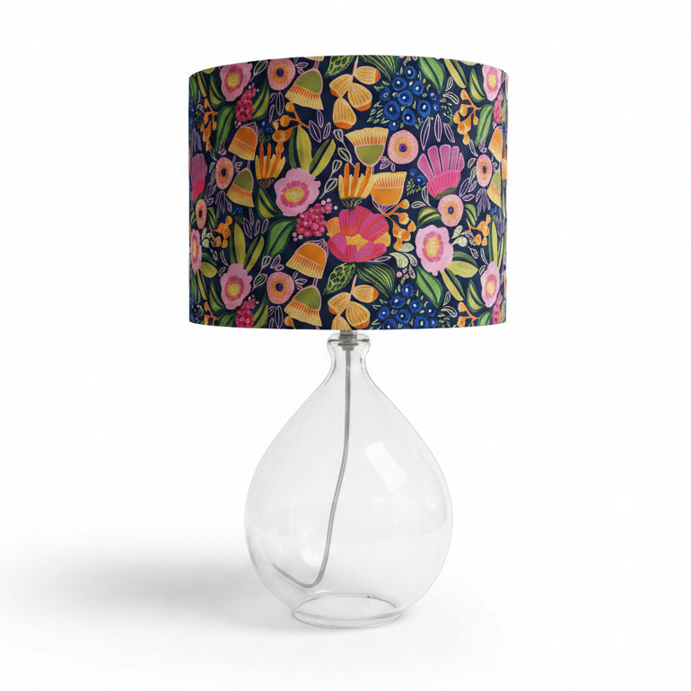 A table lamp with a colorful floral drum shade and a clear glass teardrop-shaped base, on a white background