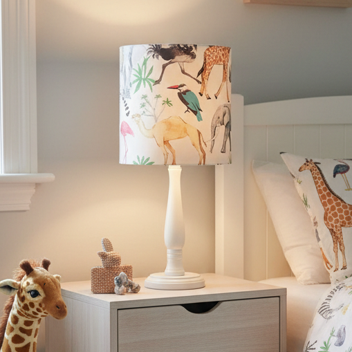 A table lamp with a cream-colored turned wood pedestal base and a cylindrical fabric shade featuring a colorful safari animal print with a giraffe, elephant, camel, bird, and palm trees on a light background.