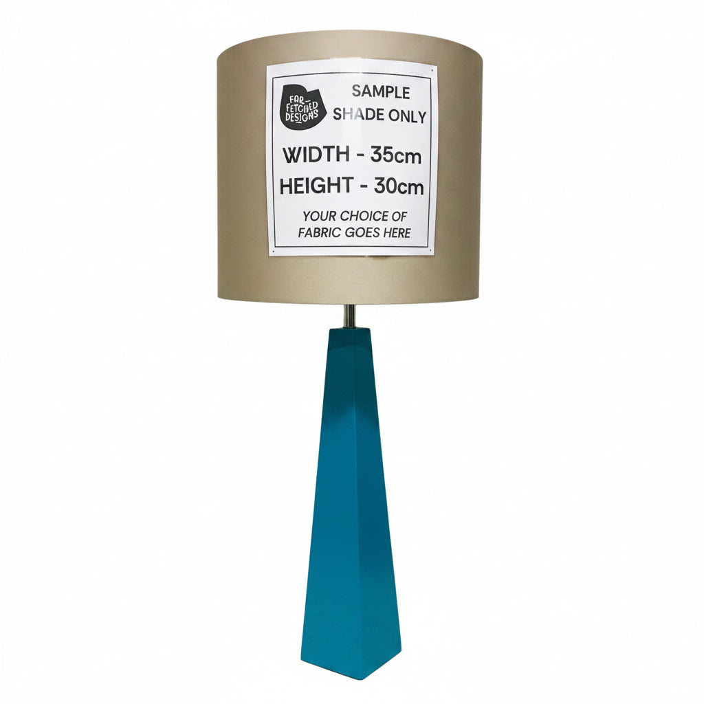 A table lamp sample with a taupe fabric shade and a teal metal base, shown against an off-white wall. A white paper label on the shade reads 'SAMPLE SHADE ONLY' with dimensions and fabric placeholder text.