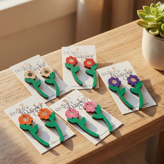 Four pairs of wooden flower-shaped earrings on white display cards, featuring green stems and leaves with red, orange, pink, and purple flowers. The cards are branded 'Rhiley Design' and laid out on a beige woven mat background.