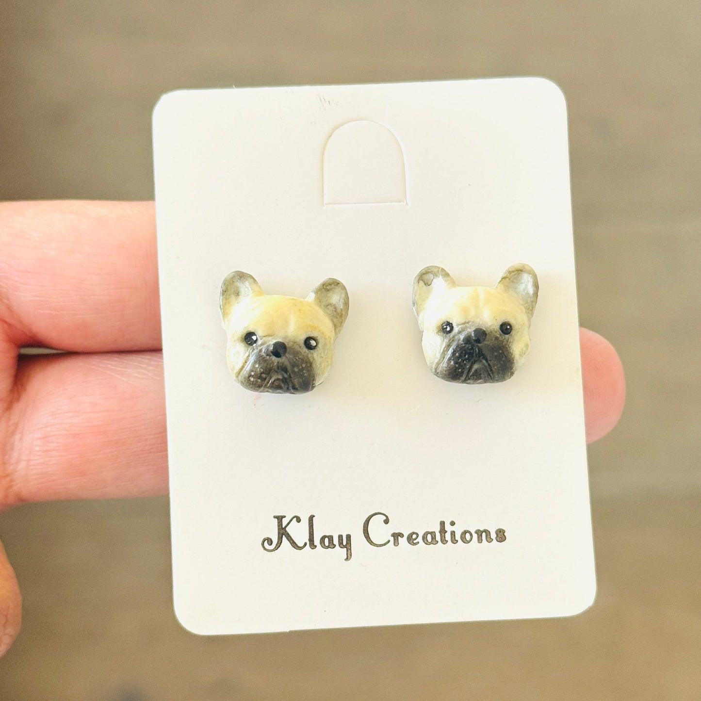 A pair of small stud earrings shaped like dog faces mounted on a white display card held by a hand against a blurred wooden background. The card is branded 'Klay Creations'.