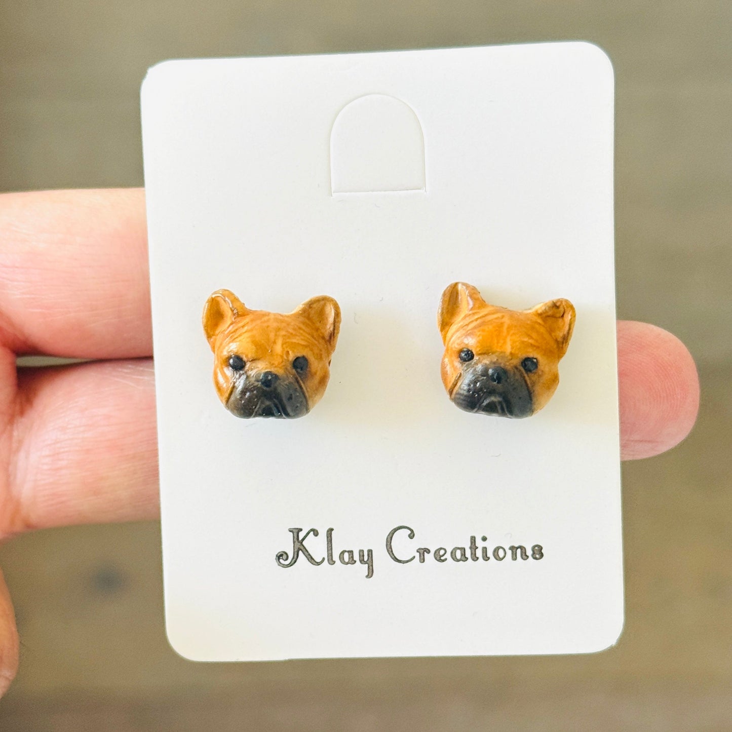 A pair of small dog-face stud earrings mounted on a white display card held by a hand. The card is branded 'Klay Creations' and photographed against a softly blurred brown background.
