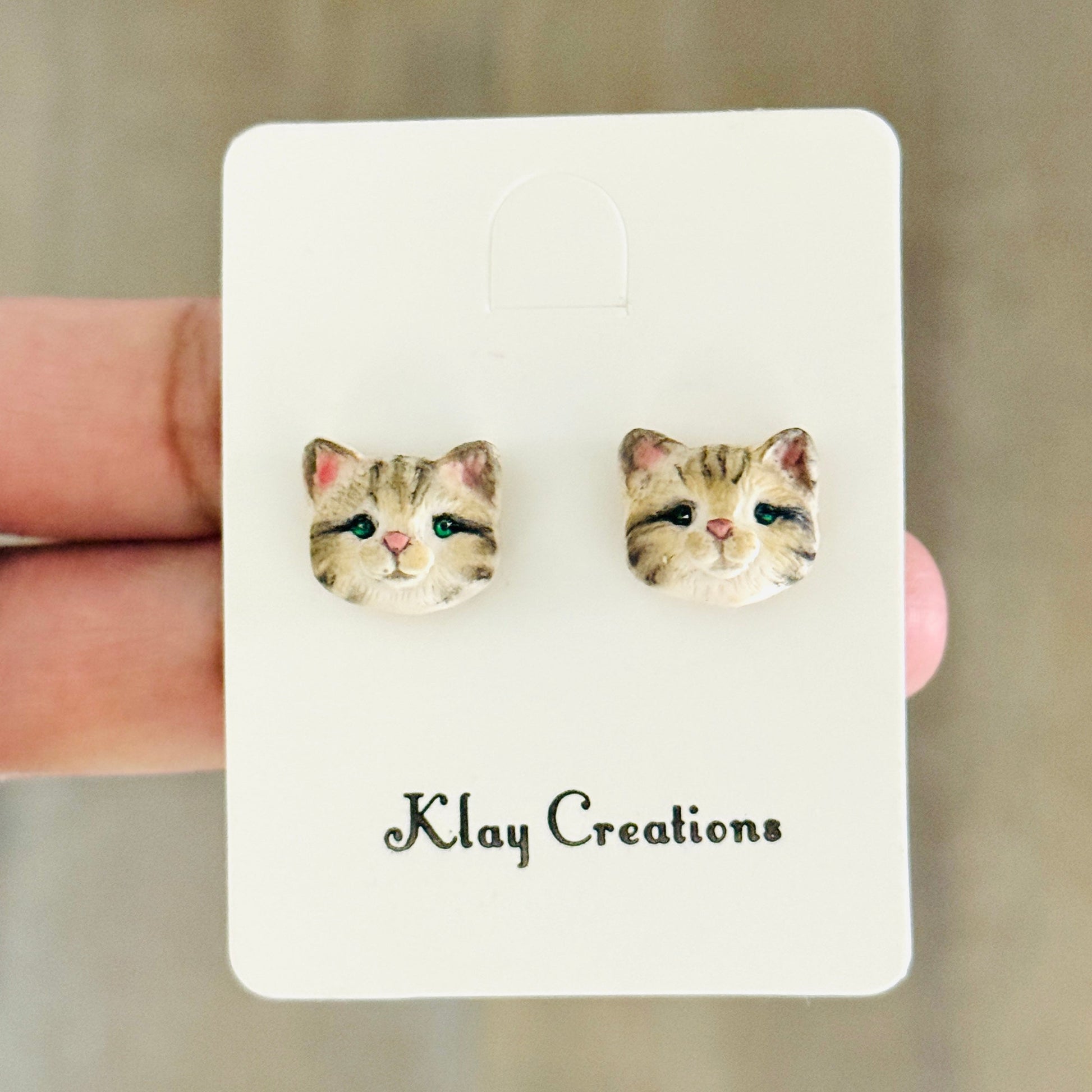 A pair of small stud earrings shaped like cat faces, displayed on a white card labeled 'Klay Creations' held by a hand against a blurred wood background.