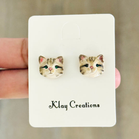 A pair of small stud earrings shaped like cat faces, displayed on a white card labeled 'Klay Creations' held by a hand against a blurred wood background.