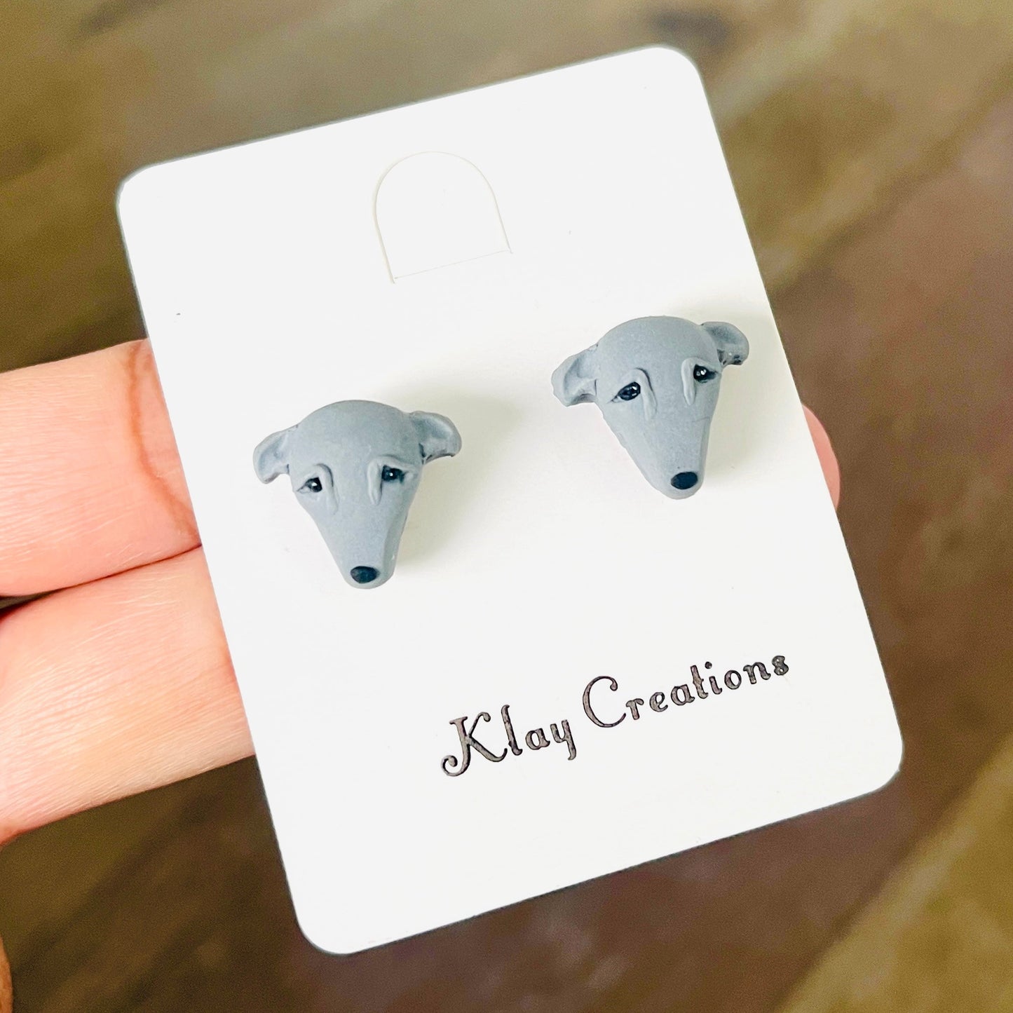 A pair of small gray stud earrings shaped like dog faces, attached to a white display card printed with the brand name 'Klay Creations'. The card is being held by a person's fingers against a blurred brown wooden background.