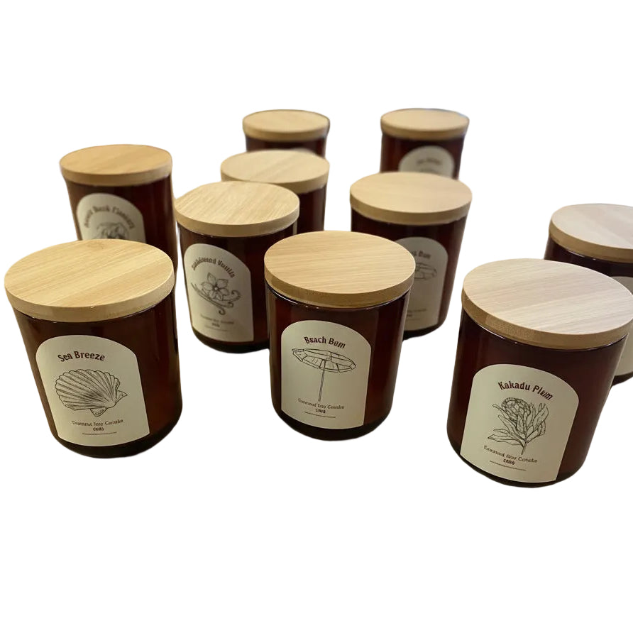 Several amber glass candle jars with light wood lids and white labels are arranged on a wooden table inside a gift shop. The labels show botanical illustrations and scent names including Sea Breeze, Quick Run, and Kakadu Plum.