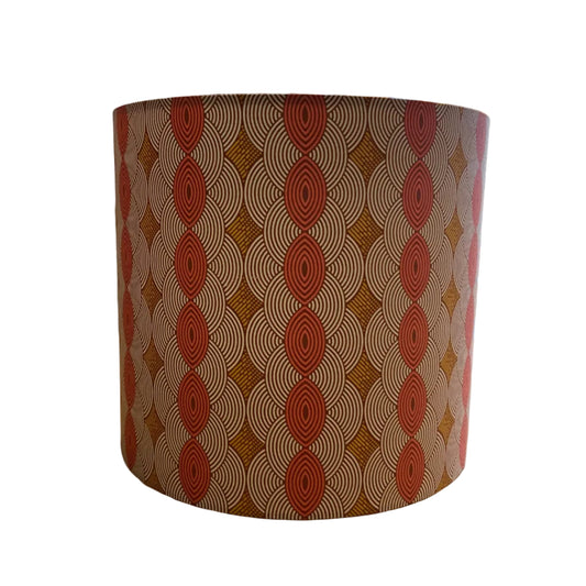A cylindrical lampshade featuring a repeating geometric pattern of red-orange concentric ovals and diamond shapes over a warm brown and beige background, shown against a plain white background.
