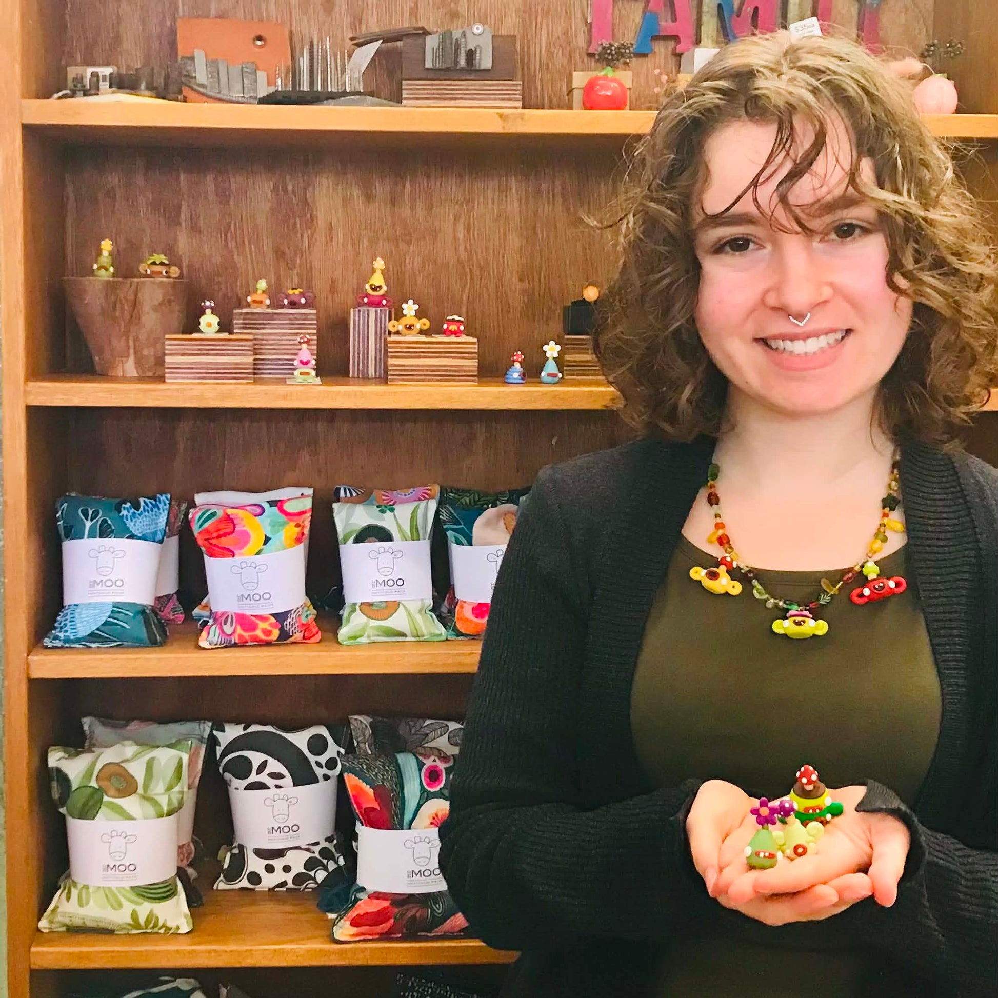 A smiling woman with curly brown hair stands in front of wooden shelves filled with colorful packaged goods and small figurines, holding a small colorful object in her hands. She wears a dark cardigan over an olive top and a chunky beaded necklace.