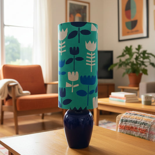 A table lamp with a glossy dark blue ceramic base and a cylindrical teal shade featuring a white and blue floral pattern, set in a cozy lounge room