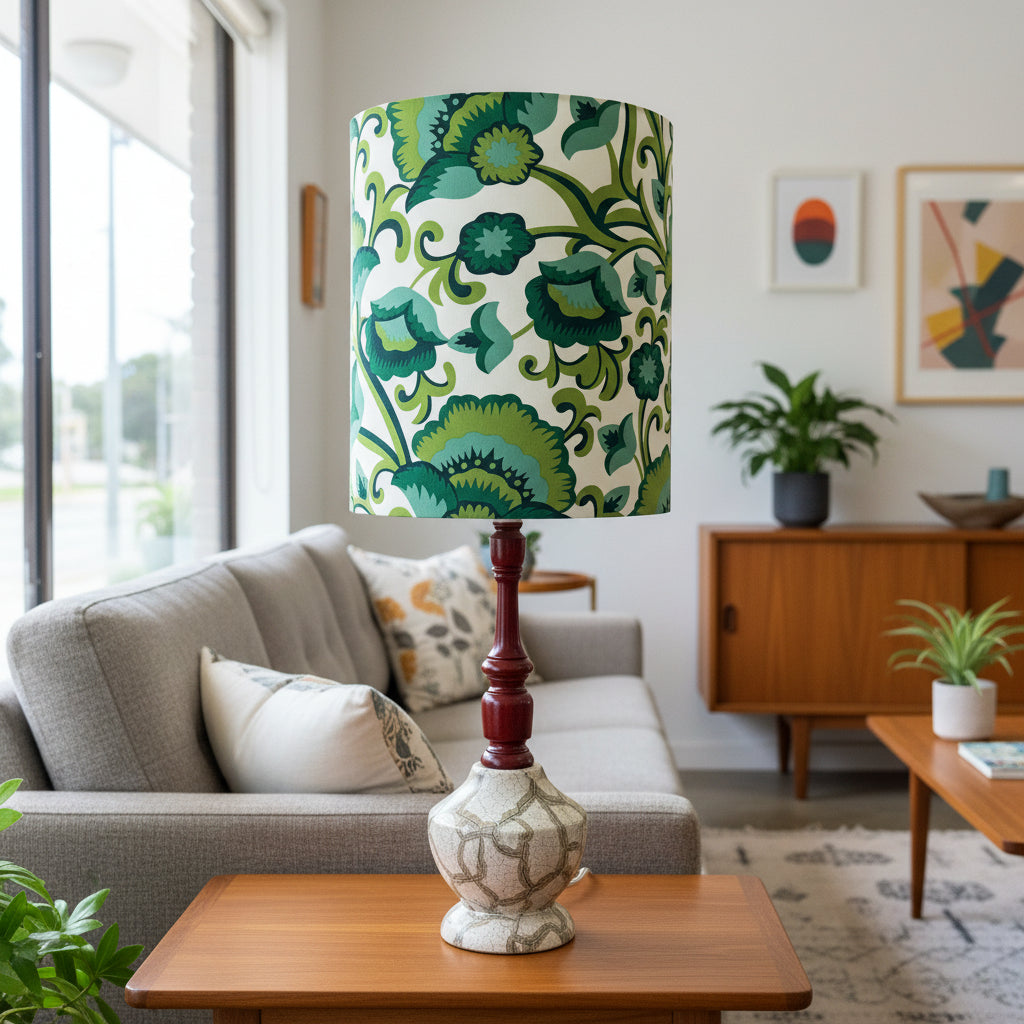 A table lamp with a green and blue floral patterned drum shade, a dark reddish-brown turned wood stem, and a textured cream ceramic base, styled on a wooden table in a bright, modern living room.