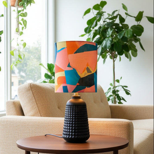 A table lamp with a textured black base and a colorful abstract geometric shade in orange, pink, blue, and yellow, styled on a wooden side table in a living room setting.