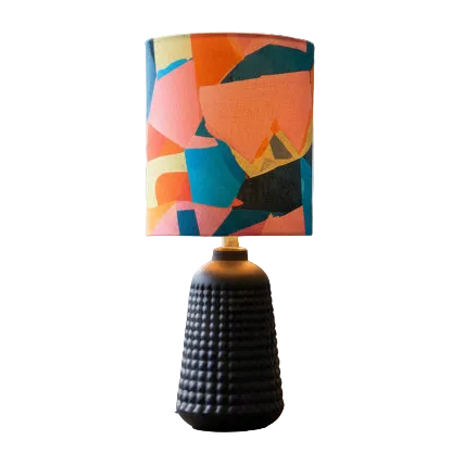 A table lamp with a textured black ribbed base and a cylindrical drum shade featuring a colorful abstract geometric pattern in orange, pink, teal, and cream, isolated on a white background.