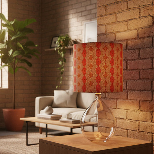 A table lamp with a cylindrical drum shade featuring a red and orange geometric pattern on a beige background, mounted on a clear glass teardrop-shaped base. The lamp is shown in a warm, modern living room setting with a brick wall, a sofa, and a potted plant.