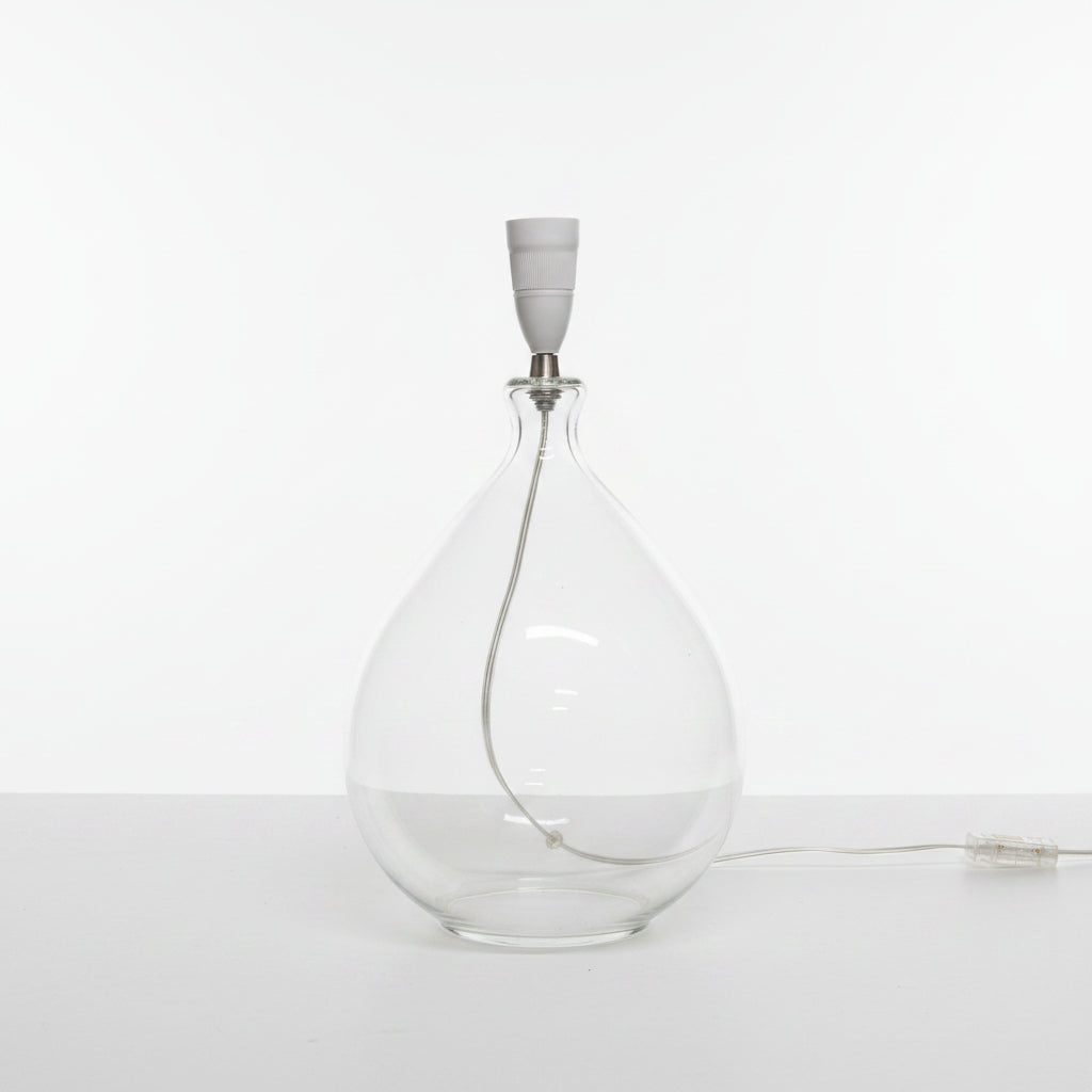 Clear glass bottle shaped lamp base with a white cap on a light-colored floor against a plain wall.