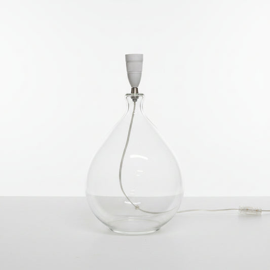 Clear glass bottle shaped lamp base with a white cap on a light-colored floor against a plain wall.