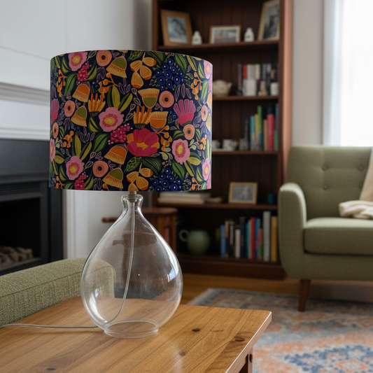 A table lamp with a colorful floral patterned drum shade and a clear teardrop-shaped glass base, placed on a wooden table in a cozy living room setting.