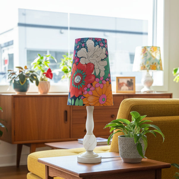 A table lamp with a white turned base and a colorful floral drum shade stands on a wooden side table in a bright living room. The shade features large red, orange, pink, and white flowers with green leaves on a teal background.