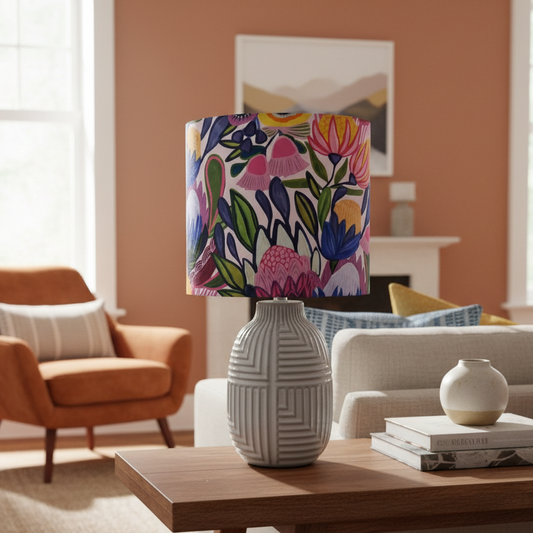 A table lamp with a white geometric ceramic base and a colorful floral drum shade, styled on a wooden coffee table in a warm, modern living room.
