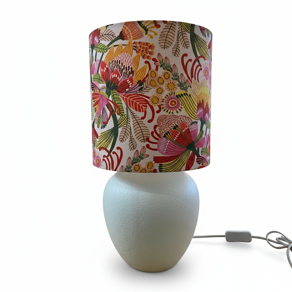 A table lamp with a textured white ceramic base and a cylindrical drum shade featuring a vibrant, multicolored floral and botanical print, set on a white background