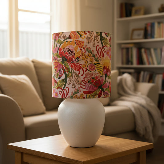 A table lamp with a textured white ceramic base and a cylindrical drum shade featuring a vibrant, multicolored floral and botanical print, set in a cozy lounge room