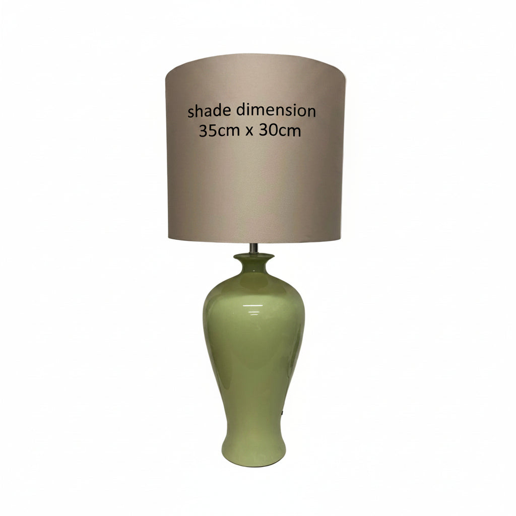 A table lamp with a glossy light green ceramic base and a large taupe-beige fabric drum shade, standing on a white surface against a white wall.