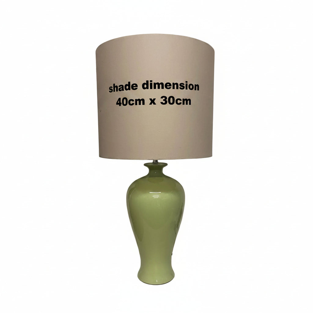A table lamp with a glossy light green ceramic base and a large beige fabric drum shade, sitting on a white table against an off-white wall.