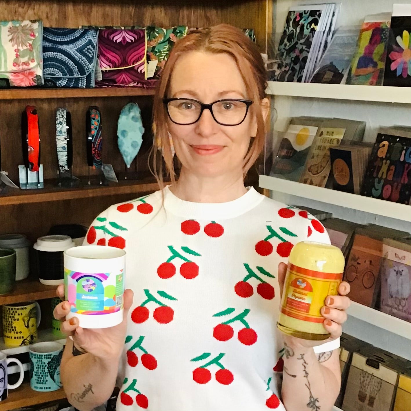 Person wearing a cherry-patterned sweater holding a mug and a jar in a store setting.