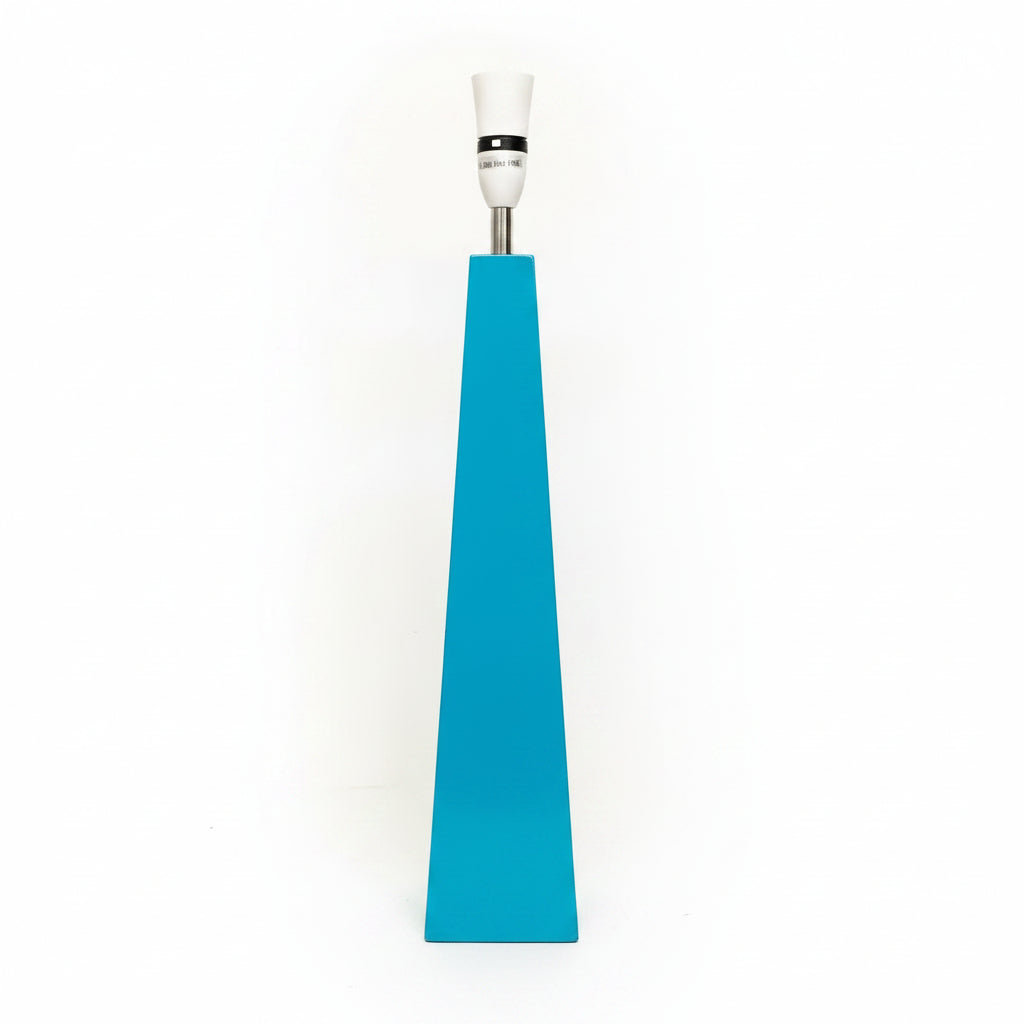 A modern geometric table lamp with a bright turquoise blue pyramid-shaped base, a silver metal stem, and a white ceramic lamp socket. A white power cord is visible on the floor next to the lamp, set against a plain off-white wall.