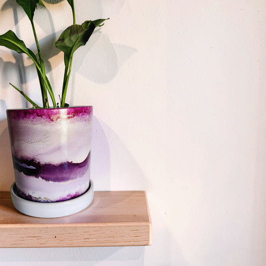 A small potted plant with green leaves sits on a light wood shelf against a white wall. The planter has a marbled ceramic design with purple, white, and pink tones, and a white saucer underneath.