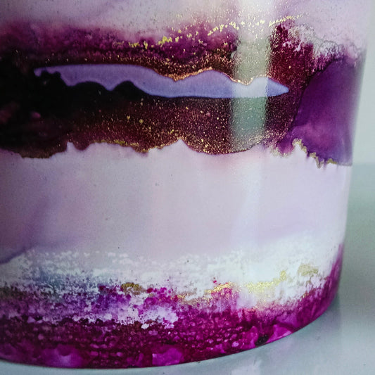 A close-up of a cylindrical container or cup with a marbled resin design featuring layers of light purple, deep magenta, white, and gold glitter.
